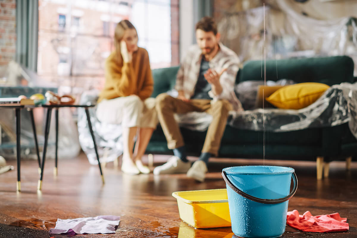 couple sitting on a couch while water drips from ceiling into a bucket